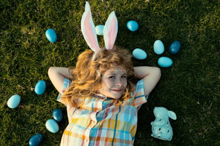 Photo for Easter bunny child hunting eggs outdoors Happy Easter day, Kid boy laying on grass in park. Top view kids face with rabbit ears - Royalty Free Image