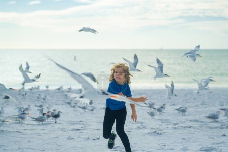 Foto de Carefree child. Happy child running with seagull birds, having fun on the beach on a hot summer day. Summer holiday vacation - Imagen libre de derechos