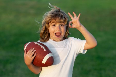Téléchargez les photos : Les enfants et le sport. Un jeune garçon jouant au football américain. Un gamin jouant avec une balle de rugby dans le parc. Enfant tenant un ballon de rugby tout en jouant au football américain dans le parc d'été - en image libre de droit