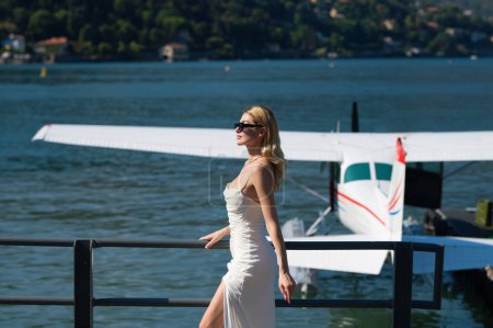 Téléchargez les photos : Vacances d'été sur le lac de Côme Italie. Femme élégante en robe tendance marchant dans la rue de la ville européenne. Femme sexy posant dans la rue près de jet plane - en image libre de droit