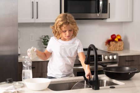Foto de A little cute boy washing dishes near sink in kitchen. Clean washed dishes, dishwashing liquid with foam - Imagen libre de derechos