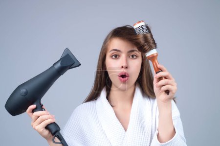 Photo for Woman in bathrobe combing hair, drying hair with hairdryer. Portrait of female model with a comb brushing hair. Girl with hairs brush and blow dryer. Hair care and beauty. Morning routine - Royalty Free Image