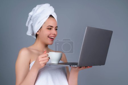 Foto de Mañana freelancer trabajando en el ordenador portátil. Mujer de negocios con toalla en la cabeza con café trabajando en el ordenador portátil. Café matutino. Retrato de mujer sonriente con taza de café usando laptop en estudio gris - Imagen libre de derechos