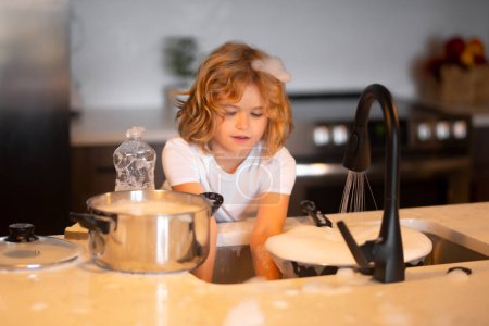 Foto de Child boy washing dishes in the kitchen interior. Cleaning dishwashing during housework. Child helping his parents with housework - Imagen libre de derechos