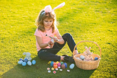 Photo for Easter kids boy in bunny ears painting easter eggs outdoor. Cute child in rabbit costume with bunny ears having fun in park - Royalty Free Image