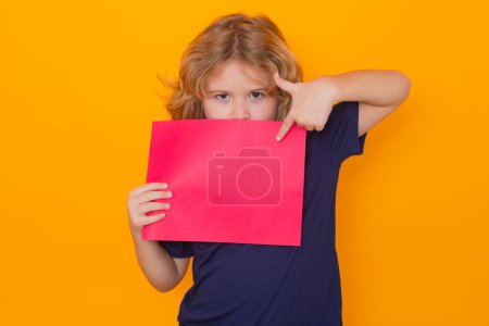Téléchargez les photos : Child pointing on empty sheet of paper, isolated on yellow background. Portrait of a kid holding a blank placard, poster. Copy space - en image libre de droit