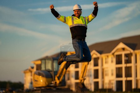 Téléchargez les photos : Jump excited worker. Builder, construction worker in helmet at construction site. Industry engineer worker in hardhat near bulldozer or digger tractor. Concept of construction industry - en image libre de droit