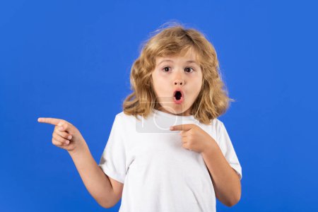 Téléchargez les photos : Excited kid boy on studio isolated background. Kid boy pointing away on blue isolated studio background. Kid with index finger pointing, copy space. Surprised face, amazed emotions of child - en image libre de droit