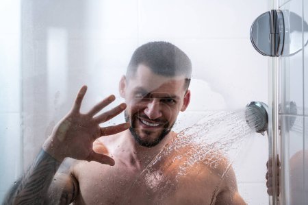 Foto de Sexy muscular man taking shower washing his body, back and shoulders. Handsome guy standing in the bathroom under hot water drops. Hygienic cleansing routine in bathroom. Skincare and bodycare - Imagen libre de derechos