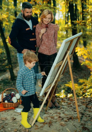 Téléchargez les photos : Les parents regardent les fils peindre. Peinture d'enfant sur toile au magnifique parc forestier doré d'automne. Concept d'art. Peinture dans la nature. Concept de créativité des enfants. Concept de famille heureuse - en image libre de droit
