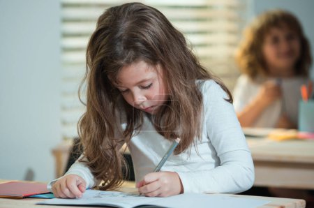 Téléchargez les photos : Écolière concentrée assise au bureau et écrivant dans un cahier d'exercices avec un camarade assis derrière - en image libre de droit