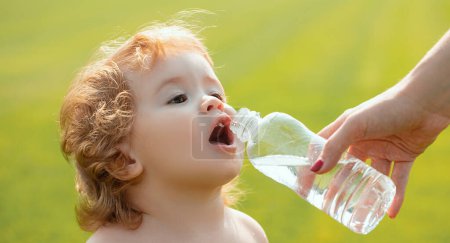 Photo for Baby drinking water from mother hands. Child drinks water from a bottle while walking on grass field, baby health. The mother gives the child a bottle of water - Royalty Free Image