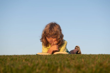 Foto de Niño inteligente leyendo libro en el parque al aire libre. Un joven chico rubio yace en la hierba verde al aire libre leyendo un libro blanco en verano - Imagen libre de derechos