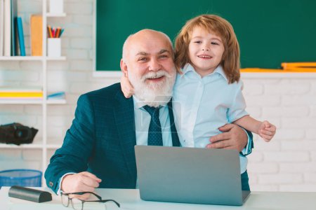 Photo for Online education. Teacher with pupil in classroom. Child with laptop. Kid is learning in class with senior mature teacher on background of blackboard - Royalty Free Image