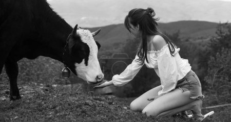 Foto de Mujer feliz alimentando vaca. Chica alimentando vaca negra y blanca en el prado. Vegano, concepto vegetariano. Toma vegetales. Estilo de vida saludable, leche de campo orgánica - Imagen libre de derechos