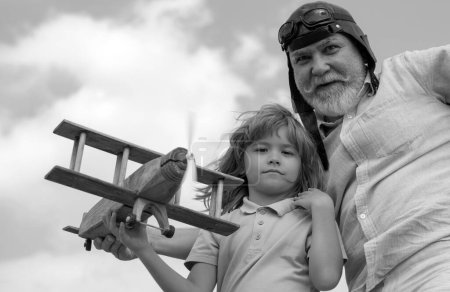 Téléchargez les photos : Enfant garçon et grand-père jouant avec un avion en bois sur fond de ciel d'été. Enfant garçon avec des rêves de voler ou de voyager. Relations familiales Grand-père et enfant - en image libre de droit