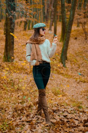 Foto de Elegante femme fatale con ropa de moda y fuma un cigarrillo en el fondo del bosque de otoño. Mujer sexy caminando en la naturaleza de otoño en traje elegante otoñal - Imagen libre de derechos