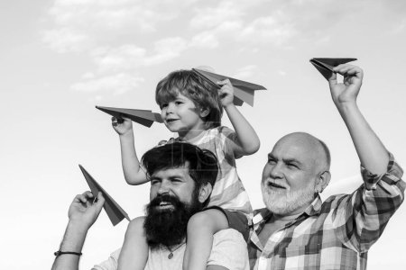 Photo for Kids playing with simple paper planes on sunny day. Father giving son ride on back in park. Generation concept. Child happy. Three men generation - Royalty Free Image