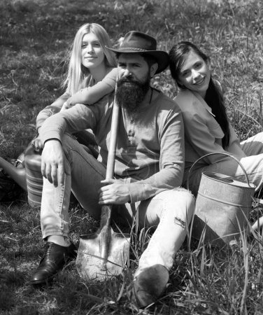 Photo for Friends sitting on grass at spring garden. Portrait of caucasian male farmer with shovel with two friend girl. Relaxing after hard work - Royalty Free Image