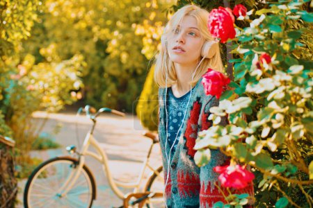 Photo for Beautiful stylish woman in autumn outfit enjoy in fall park. Girl with headphones looks dreamy. Red roses and bicycle at background. Warm september weather. Autumn concept - Royalty Free Image