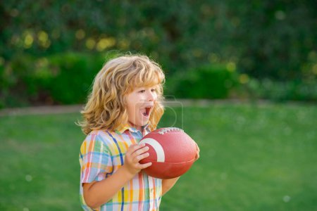Téléchargez les photos : Enfant avec ballon de rugby. Enfant mignon s'amuser et jouer au football américain sur le parc d'herbe verte - en image libre de droit