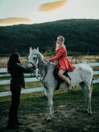 Téléchargez les photos : Femme chevauchant cheval arabe gris en robe rose. Un bel homme barbu conduisant un cheval pour une bride et regardant sa petite amie. Romantique histoire d'amour de couple sensuel - en image libre de droit