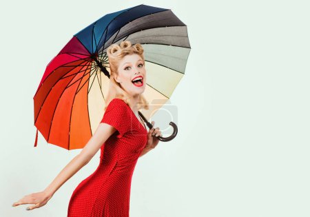 Foto de Feliz pinup mujer con paraguas en tiempo lluvioso. Lluvia arco iris otoño estado de ánimo, pin up chica en vestido rojo aislado blanco - Imagen libre de derechos