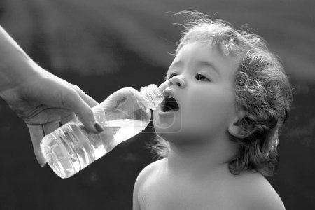 Foto de Bebé bebiendo agua de manos de madre. El niño bebe agua de un biberón mientras camina en el campo de hierba, la salud del bebé. La madre le da al niño una botella de agua. - Imagen libre de derechos