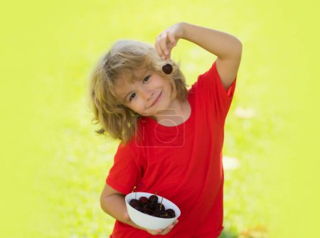 Téléchargez les photos : L'enfant en t-shirt rouge mange des cerises. Visage excité. Cerise pour les enfants. Adolescent enfant tenir cerises assiette. Expression étonnante, joyeuse et heureuse - en image libre de droit