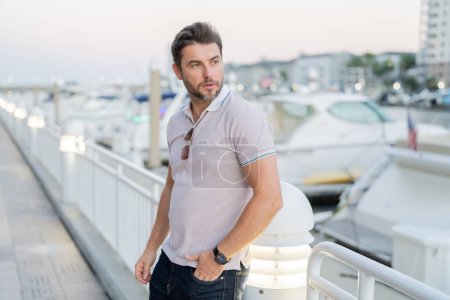 Portrait of handsome male model outdoor. Stylish man dressed in polo. Fashion male posing on the street background. Urban style. Rich man dreaming about riches near the yacht