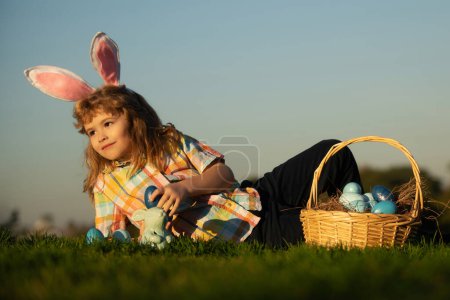 Foto de Niño lindo conejito con orejas de conejo. Niños cazando huevos de Pascua en el parque poniendo en la hierba sobre el fondo del cielo con espacio de copia - Imagen libre de derechos