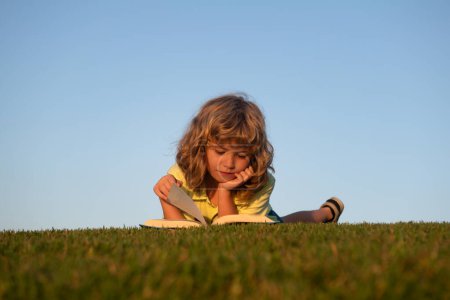 Photo for Kid reading book laying on grass on grass and sky background with copy space. Outdoor school, learning kids - Royalty Free Image