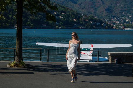 Photo for Fashion young woman in stylish dress walking near jet. Woman tourist getting out of jet airplane. Summer destination travel. Summer vacation on Como lake Italy. Travel and active lifestyle concept - Royalty Free Image