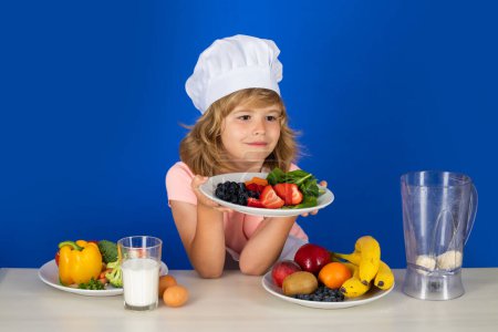 Foto de Retrato de un niño de 7, 8 años de edad en tapa de cocinero y placa de retención de delantal con frutas haciendo ensalada de frutas y cocinar alimentos en la cocina. Linda pequeña rubia feliz chef sonriente - Imagen libre de derechos
