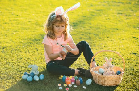 Téléchargez les photos : Enfant de Pâques garçon dans des oreilles de lapin peignant des œufs de Pâques en plein air. Enfant mignon en costume de lapin avec des oreilles de lapin s'amusant dans le parc - en image libre de droit