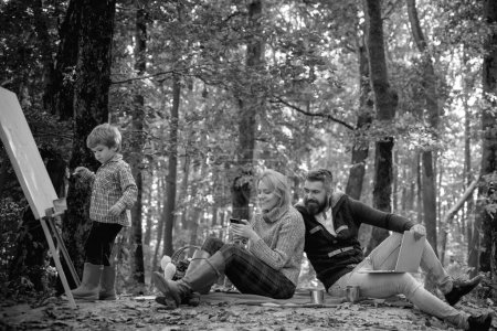 Téléchargez les photos : Une jeune famille souriante assise dans des feuilles un jour d'automne en forêt. Une famille heureuse de trois personnes passe un bon moment au parc. Petit garçon peint avec un chevalet de dessin. Concept de parents aimants - en image libre de droit
