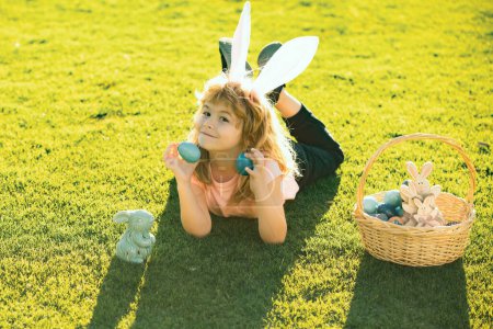 Foto de Feliz Pascua. Niño con traje de conejo con orejas de conejo cazando huevos de Pascua en la hierba en el parque de primavera - Imagen libre de derechos