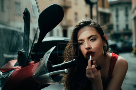 Photo for Makeup in motion. Fashion girl with red lips put lipstick looking in a mirror of motorcycle. Street fashion. City life - Royalty Free Image