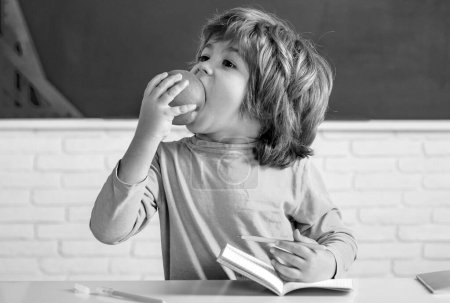 Foto de Niño amigable comer manzana en el aula cerca del escritorio de pizarra. Aula de la escuela primaria. Concepto de educación para niños - Imagen libre de derechos