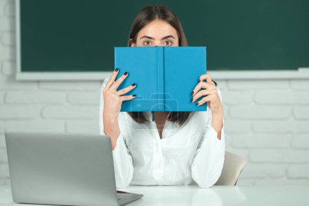 Photo for Beautiful student, cute young woman in class at school. Female college student hiding behind an open book and looking away. Girl covering face with book - Royalty Free Image