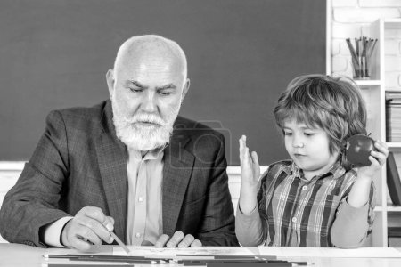 Photo for Portrait of senior teacher and little boy kid sitting at desk in classroom. Pupil and Teacher in classroom. Senior teacher in classroom with elementary school kid - Royalty Free Image