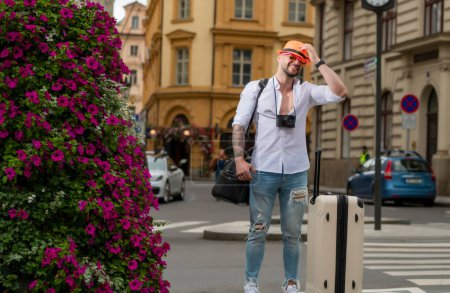Foto de Hombre turista viajero en ropa casual sombrero con maleta. Hombre viajero con bolsa de viaje al aire libre. Retrato de hombre joven caucásico en sombrero y gafas de sol en las vacaciones de verano - Imagen libre de derechos