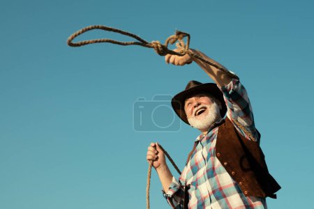 Foto de Viejo vaquero occidental con cuerda de lazo. Barbudo hombre del oeste salvaje con chaqueta marrón y sombrero - Imagen libre de derechos
