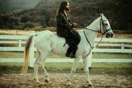 Foto de Hombre barbudo guapo montando a caballo en la granja. Hermoso caballo con jinete hombre trotando en el campo otoñal. Concepto de amor ecuestre y animal. Jinete en caballo árabe gris en el campo - Imagen libre de derechos
