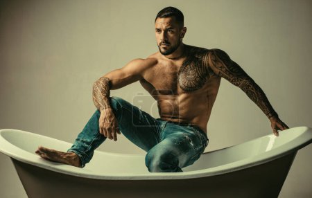 Homme macho sexuel dans le bain. Homme tatoué musclé fort tenant bouteille de champagne et posant dans la salle de bain. Beau barbu homme torse nu en jeans avec corps sexy dans la salle de bain
