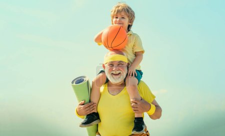 Foto de Sportsman abuelo y niño sano con pelota de baloncesto sobre fondo azul cielo. Abuelo e hijo haciendo ejercicios. Joven - viejo entrenamiento - Imagen libre de derechos