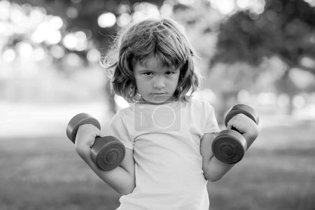 Photo for Strong child boy is doing exercises with dumbbells in summer park - Royalty Free Image
