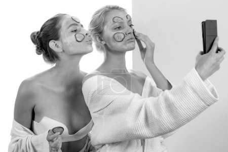 Photo for Smiling happy girls with natural clay mud mask and fresh slices of cucumber at face are having fun and taking selfies. Good mood, friendship and healthy skincare routine - Royalty Free Image