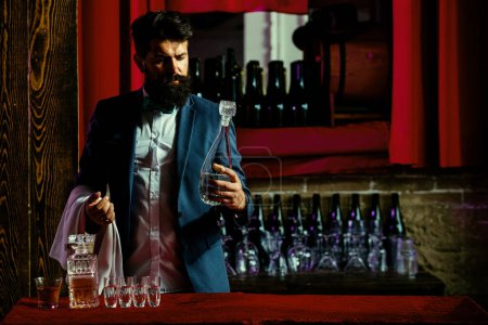 Téléchargez les photos : Beau barman à longue barbe et moustache au visage sérieux fait cocktail alcoolisé en cuir daim vintage sur fond de bar rouge violet - en image libre de droit