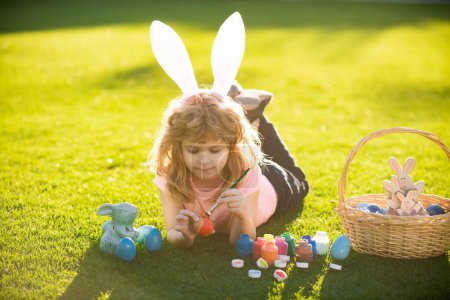 Téléchargez les photos : Enfant pondant sur l'herbe dans les oeufs de Pâques parc esprit. Des enfants lapins de Pâques. Enfant garçon en lapin Oreilles de Pâques peinture oeufs - en image libre de droit